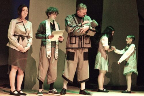 Scene from Directing Memory, Style, and Dystopian: The Giver at Gaslight Baker Theatre Mom in black piped asymmetric shirt and skirt, Jonas in slanted pants and almost star trek like shirt, Dad in block and outlined jacket, Fiona in school uniform with asymetric hem and slanted button line. Lily in original v-hem pinafore style dress.