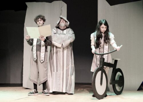 coummunity theatre Theatrical production of the Giver with Fiona on a stylized vintage vibe bike, Jonas looking at a folder and Giver looking over his shoulder.